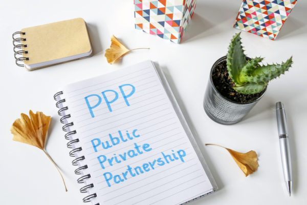 PPP Public Private Partnership geschrieben in Notizbuch auf weißem Hintergrund|Die Wörter Private vs Public Wörter auf Papierpfeilen geschrieben auf Kork Bord festgesteckt.