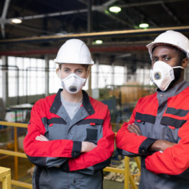 Portrait of serious workers in respiratory masks and hardhats standing with crossed arms on frame bridge in factory shop