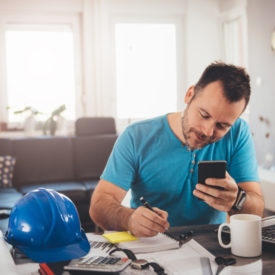 Man in blue shirt writing notes and holding smart phone at home office