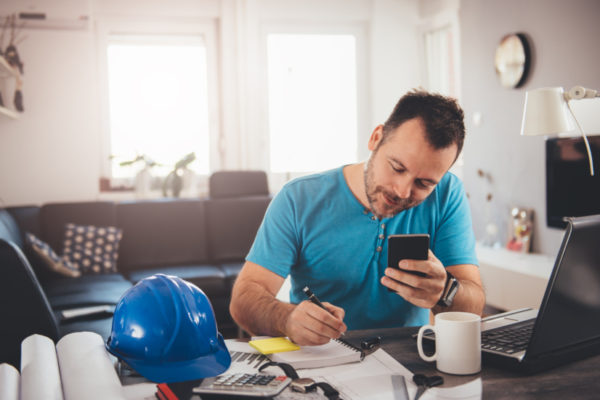 Man in blue shirt writing notes and holding smart phone at home office