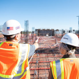 A site manager and trainee site manager in hard hats and high visibility vests look out over a construction site.