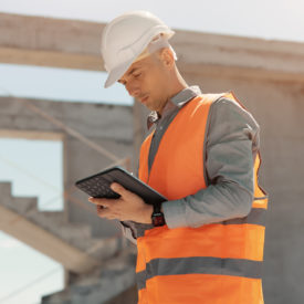 An engineer in an orange vest and white construction control helmet conducts an inspection with a tablet in his hands.