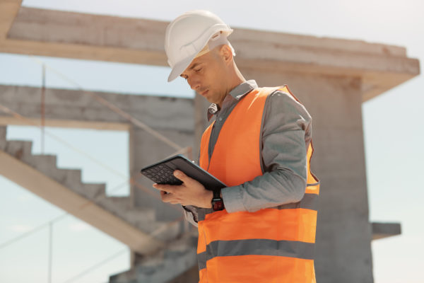 An engineer in an orange vest and white construction control helmet conducts an inspection with a tablet in his hands.